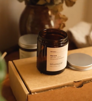 Empty small amber glass jar with a white label and black text. The jar is sitting on top of a kraft box. Next to the jar sits a silver aluminium lid. Another candle is in the background next to a dried plant in a vase.