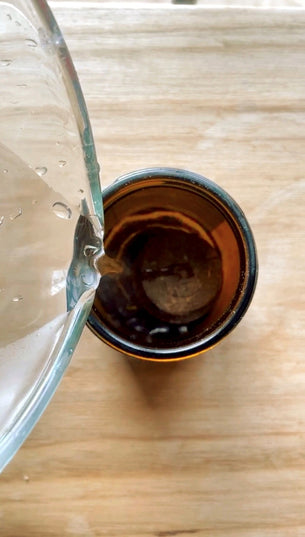 Birdseye view of a glass jug pouring water into an amber glass jar filled with soil pellets, with a wooden tabletop in the background.