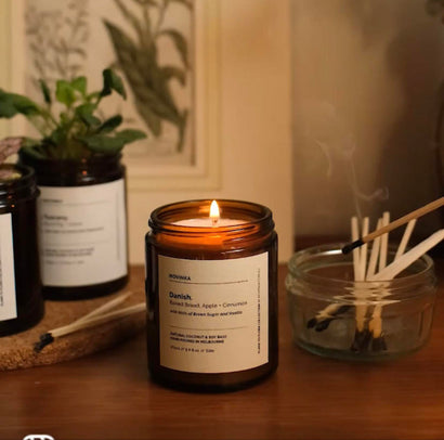 Lit small amber jar candle on a wooden table. Framed image of plants in the background. On the right, next to the candle is a glass bowl of used matches. On the left in the background, is an amber jar with a white label with a pot plant inside.